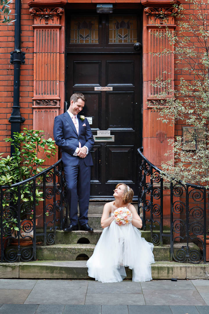 A casual and relaxed wedding portrait featuring a bride and groom. The bride is taking a pew on the steps of a classic Mayfair redbrick building with black door, resting her wedding bouquet on her knees while looking up at the Groom. The groom is casually leaning against the ornate cast iron railings.