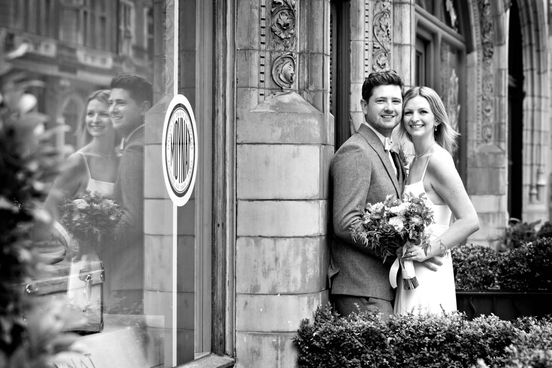 A black and white photograph of a bride and groom snuggled together. The photographer cleverly uses the glass of a shop window to capture a reflection of the bride and groom, so you see the couple twice.
